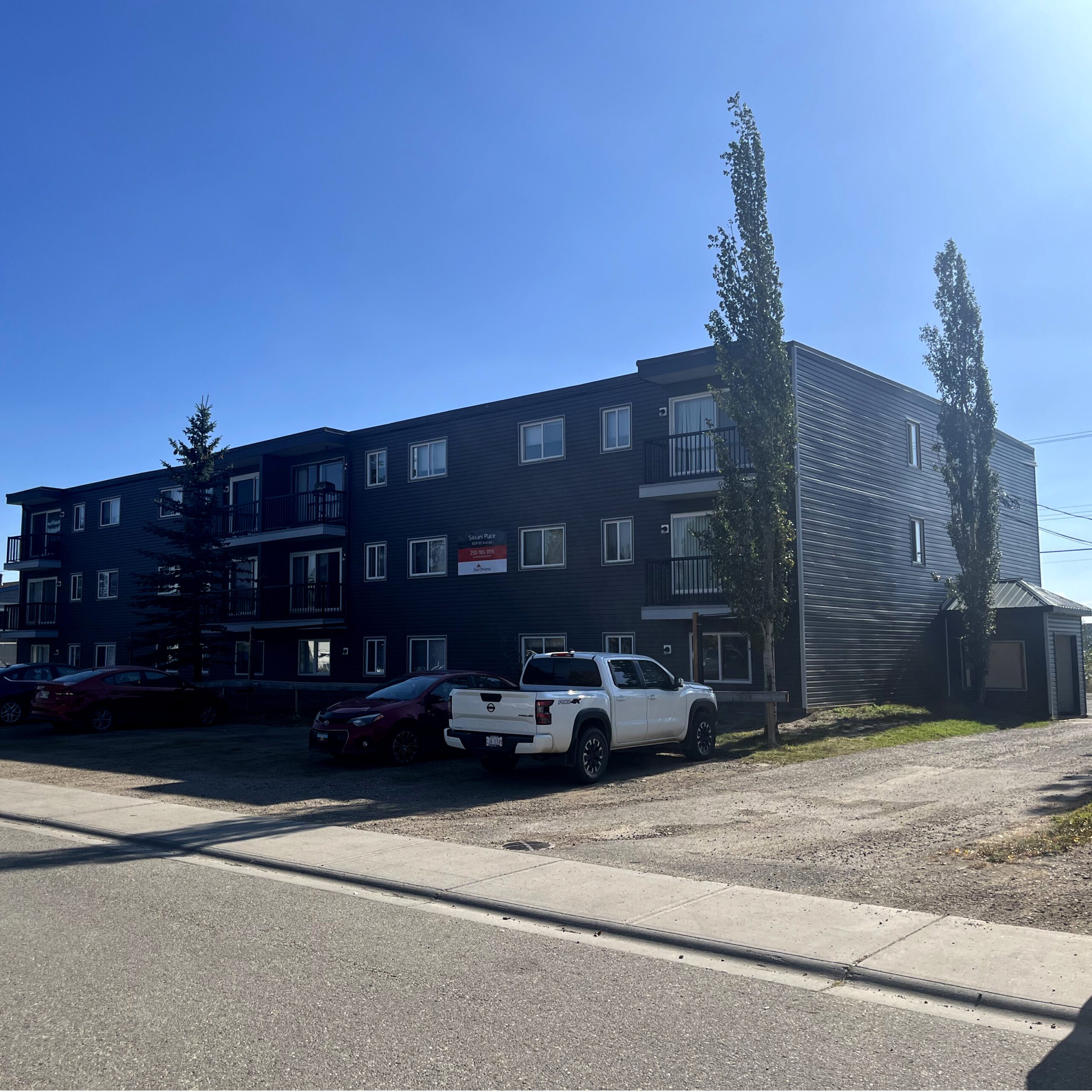 Modern residential apartment building with multiple balconies and parked vehicles on a sunny day